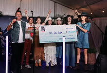 Group of women and men celebrating on stage with a large check for US Ski Team at a winter event.