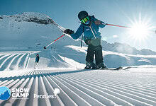 Young child in ski gear skiing on groomed snow with mountains in the background and bright sunlight overhead.