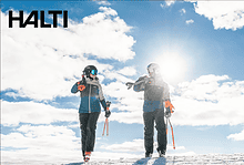 Two skiers in Halti outdoor clothing on snowy mountain, bright sun and blue sky in background.