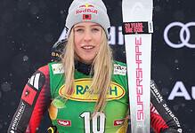 Female skier in racing gear with bib number 10, snowy backdrop, Red Bull beanie, holding a ski pole, promotional branding visible.