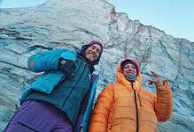 Two explorers in colorful winter jackets standing in front of a glacier, smiling and enjoying the winter outdoor adventure.