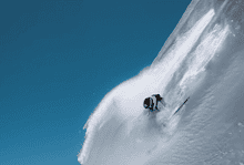 Skier descending a steep, snow-covered mountain slope during bright winter day, showcasing thrilling winter sports activity.