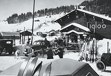 Vintage black and white winter scene at a ski resort with people, cars, and snow-covered buildings.